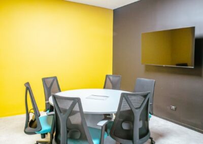 Small meeting room with a round table, office chairs, and a yellow accent wall.