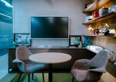 Small office meeting space with a round table, two chairs, a wall-mounted screen, and shelves.