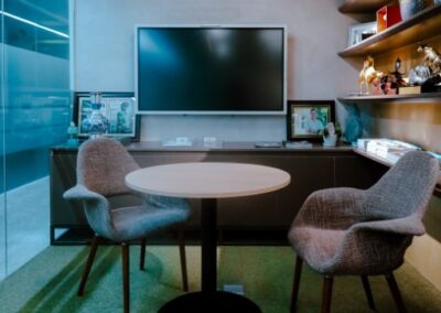 Small office meeting space with a round table, two chairs, a wall-mounted screen, and shelves.