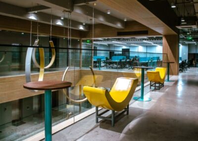 Modern office interior with glass walls, high tables, and yellow chairs along a corridor.